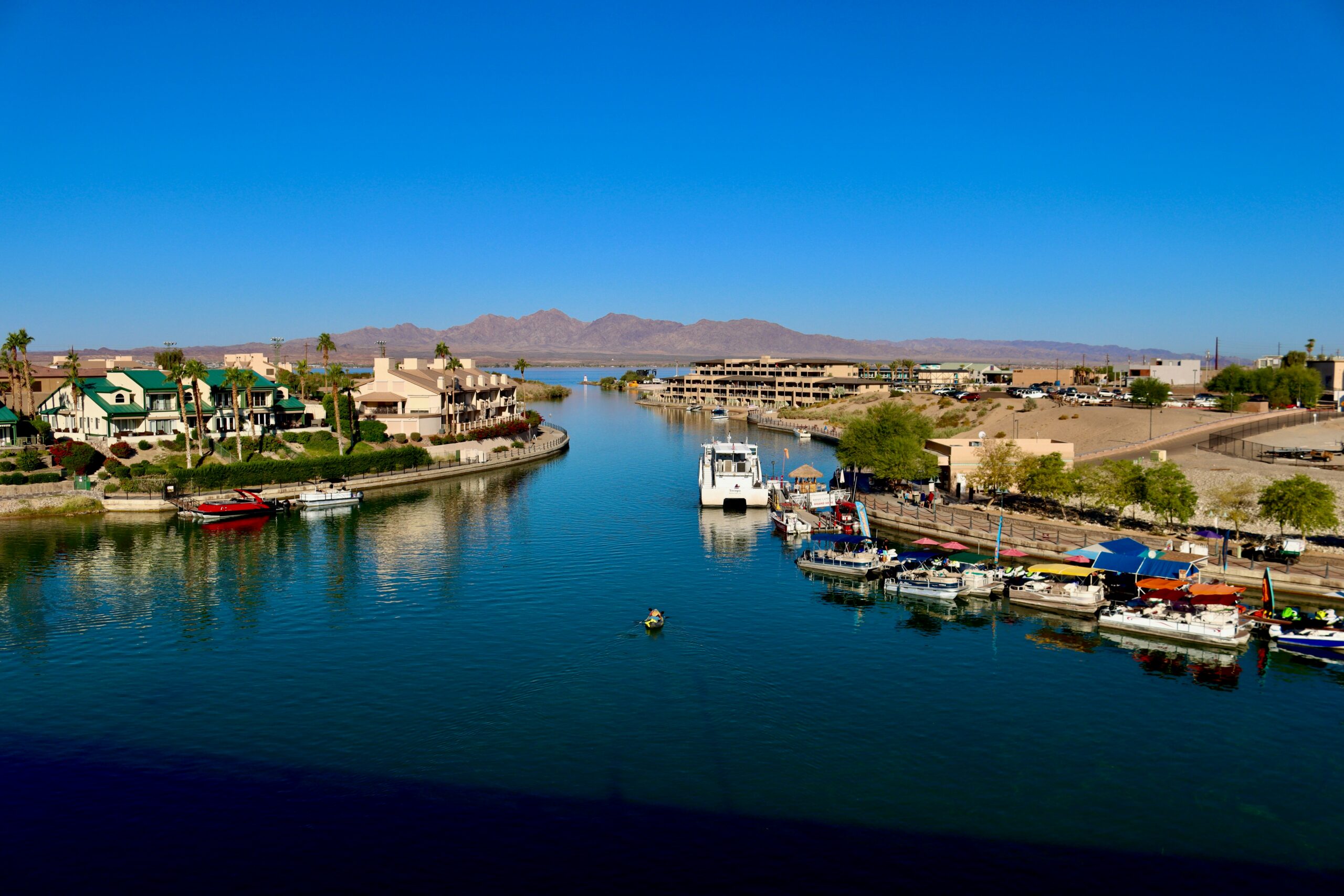 Aerial view of a waterfront resort in Lake Havasu City featuring a marina with docked boats, a canal leading to the lake, waterfront accommodations, and desert mountains under a clear blue sky